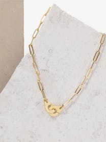 A Dinh Van gold chain link necklace with an iconic "handcuff" clasp lies on a light gray, textured surface, angled diagonally.