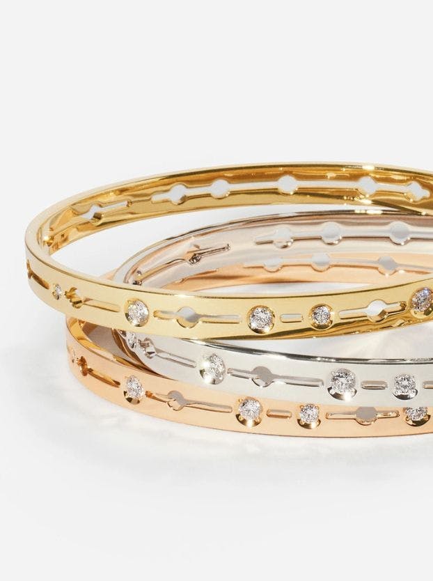 Three Dinh Van bangles are stacked, made of yellow gold, white gold, and rose gold. Each bangle has cut-out circular patterns interspersed with small, bezel-set diamonds.