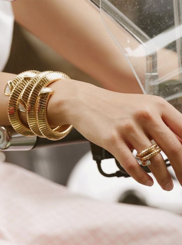 A hand on a scooter wears three wide, ridged gold bangles and a matching ridged ring with a clear stone.