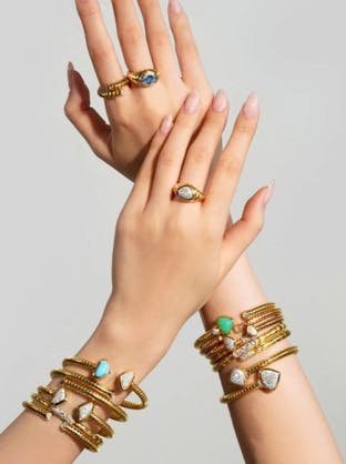 Two hands display multiple Marina B gold rings and bangles, some with colored gemstones, diamonds, and shield or teardrop shapes.