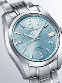A close-up of a Grand Seiko watch with a light blue, textured dial and a stainless steel bracelet, showing the date window.