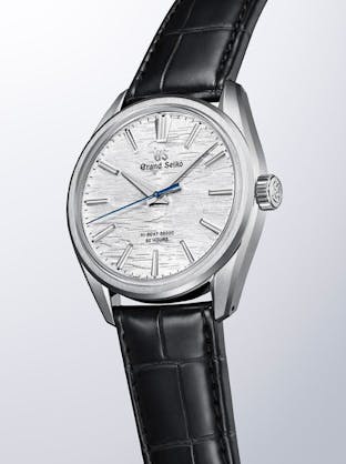 A close-up of a Grand Seiko watch with a white, textured dial, blue seconds hand, and black leather strap.