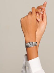 A woman's arm displays a rectangular Longines Mini DolceVita watch with a silver dial and steel bracelet, against a neutral background.