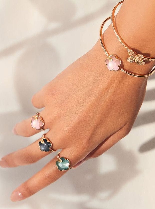 A close-up of a hand displays Pasquale Bruni floral rings with pink, dark, and green stones, alongside a thin gold bangle with a sparkly butterfly.