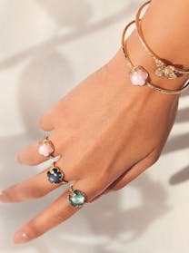 A close-up of a hand displays Pasquale Bruni floral rings with pink, dark, and green stones, alongside a thin gold bangle with a sparkly butterfly.
