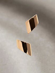 Two sleek, highly polished, rhombus-shaped rose gold earrings appear to float against a soft, subtly textured gray background, reflecting light and shadow.