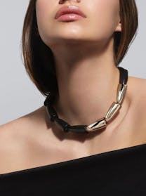A close-up view of a woman's neck and décolletage showcasing a bold necklace with alternating polished gold and matte black elongated, rounded links. She wears a black off-the-shoulder top.