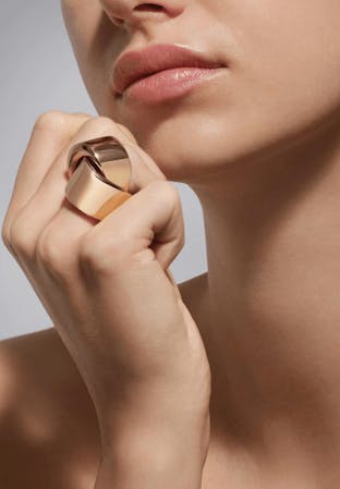 A close-up shot focuses on a woman's lower face and right hand, which is adorned with a large, sculptural rose gold ring featuring interlocking, polished geometric shapes, resting near her chin.