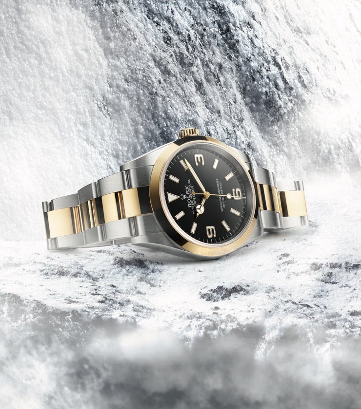 A Rolex Explorer watch with a black dial and two-tone gold and silver bracelet, displayed against a snowy, mountainous background.