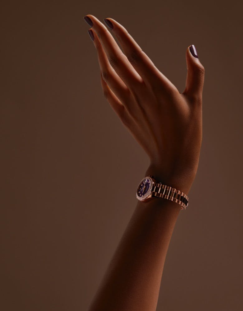 A hand with manicured nails in a dark polish wearing a Rolex Lady Datejust watch with a dark face and a gold band, set against a brown background.