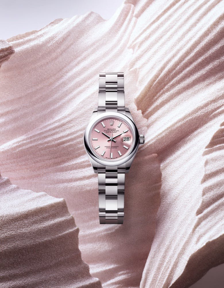 A Rolex Lady Datejust watch with a silver bracelet and pink dial, elegantly displayed against a textured, wave-like background.