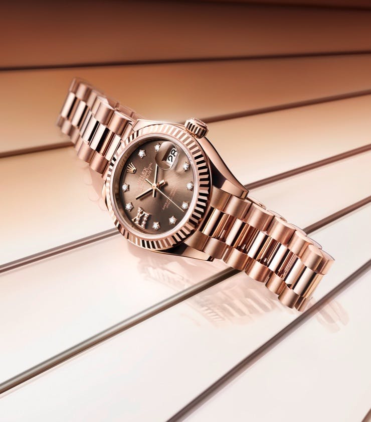 A Rolex Lady Datejust watch with a rose gold bracelet and diamond markers, elegantly positioned on a reflective, linear backdrop.