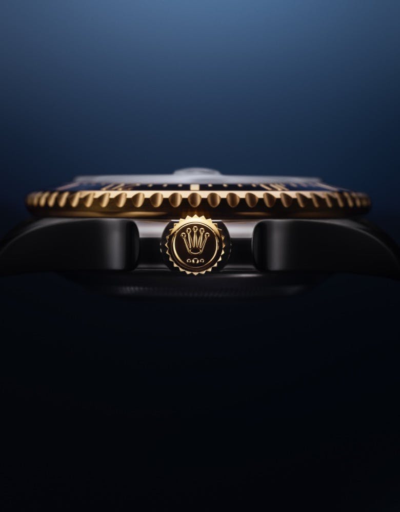 A close-up view of the Rolex Sea Dweller's crown, featuring a gold emblem and bezel, set against a dark blue background.