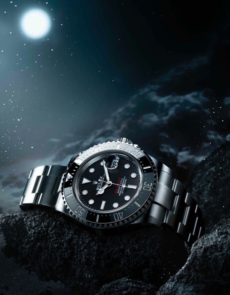 A Rolex Sea Dweller watch with a black dial and silver bracelet, displayed on a rocky surface under a moonlit sky with clouds and stars in the background.