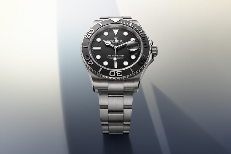 A Rolex Yacht-Master watch with a black dial and silver bracelet, displayed against a gradient background with shades of light and dark gray.