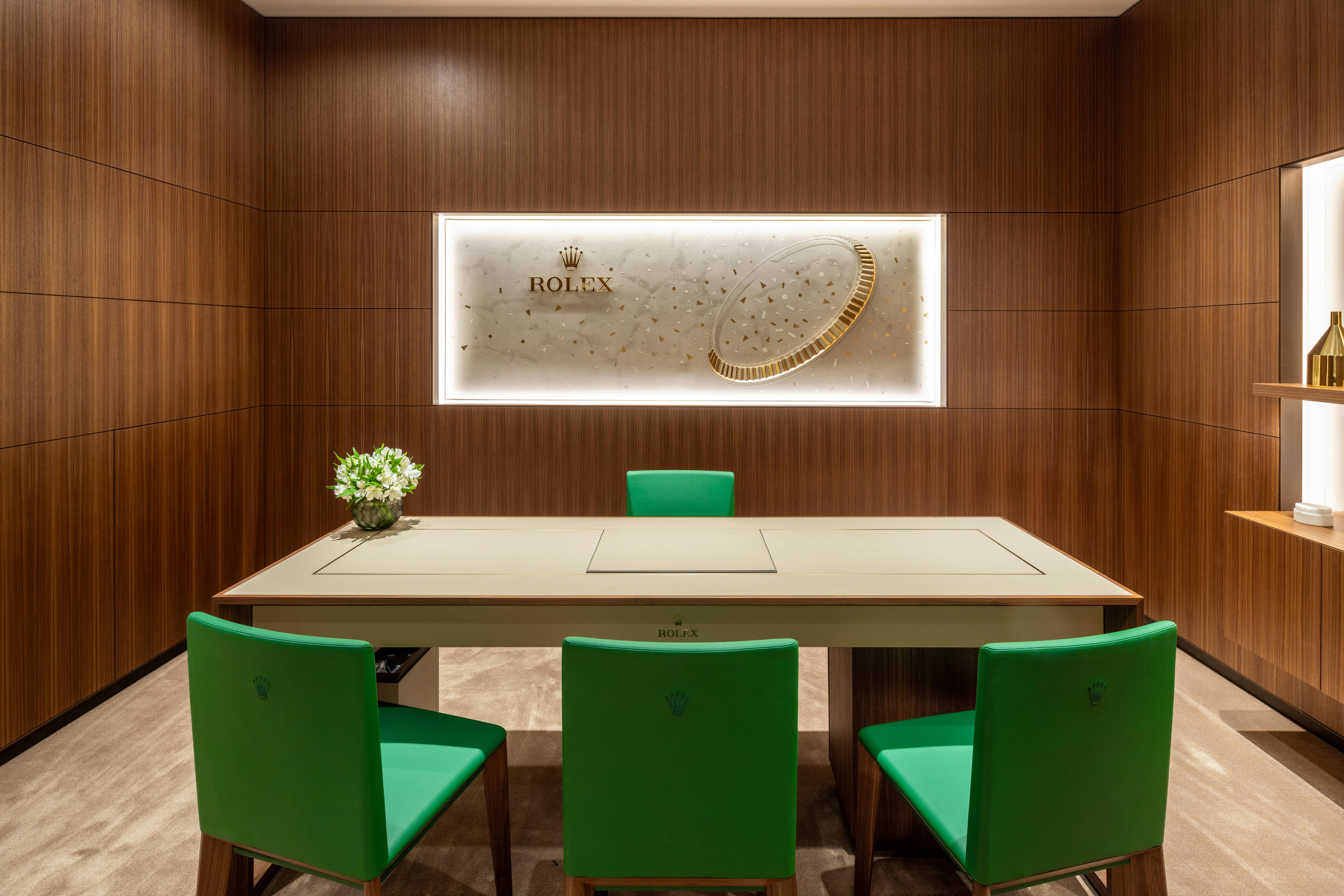 A private room in a Rolex showroom featuring a wooden interior, green chairs, a central table, and an illuminated Rolex sign on the wall.